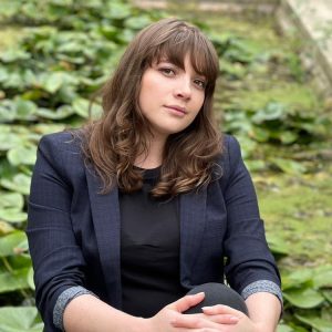 A woman with brunette, slightly curly hair and brown eyes. She wears dark navy blue blazer.