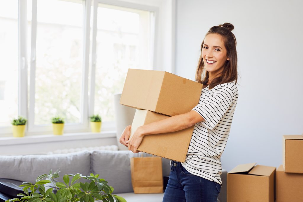 Smiling woman packing boxes for storage during her home move. There are cardboard boxes and a living room on the background.